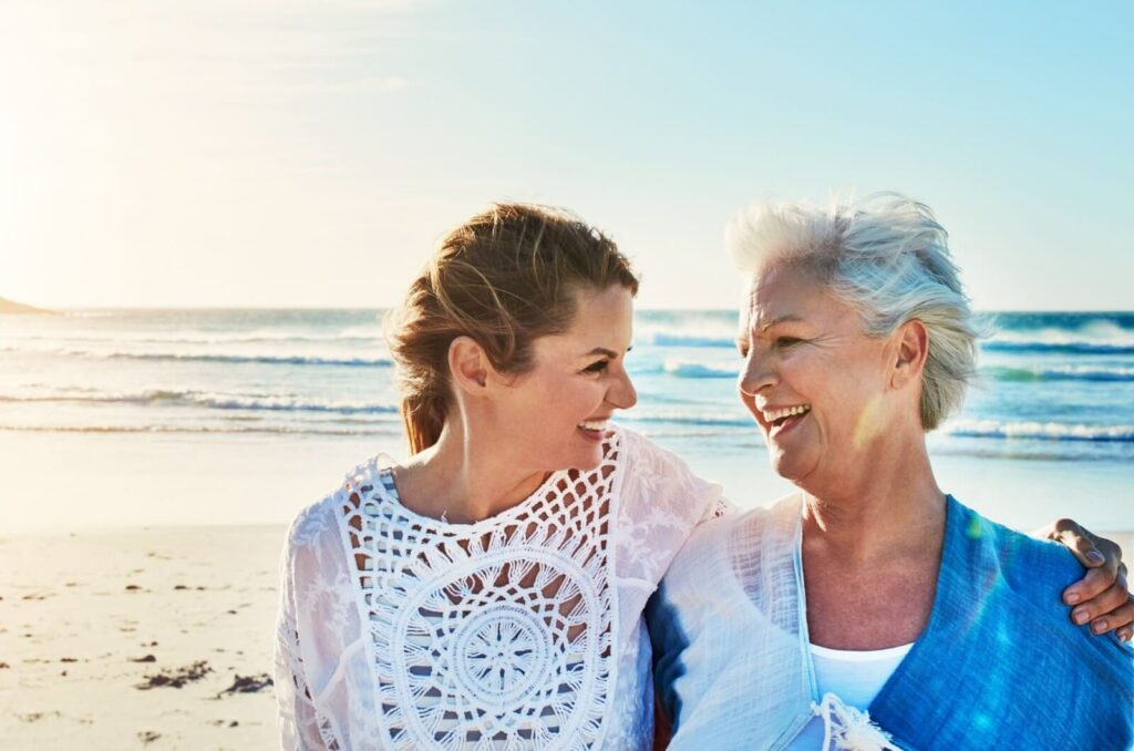 Adult daughter and  mother standing on the beach in the wind and smiling at each other while hugging from the side.