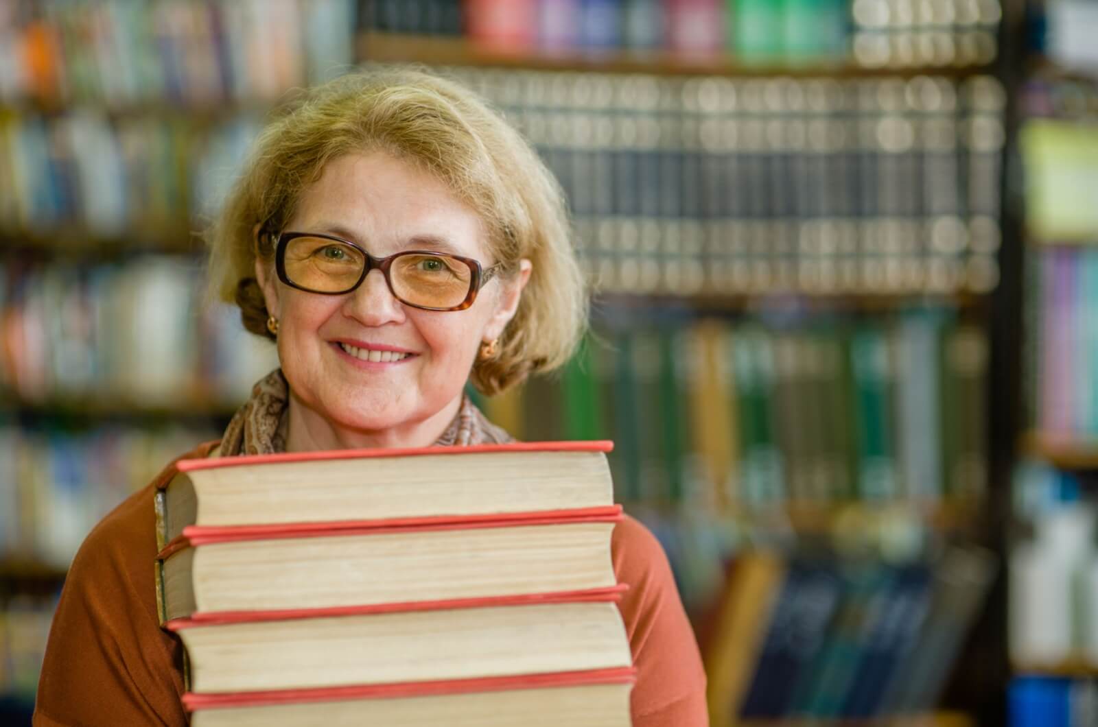 An older adult in the library, smiling and holding a stack of hardcover books for their book club.