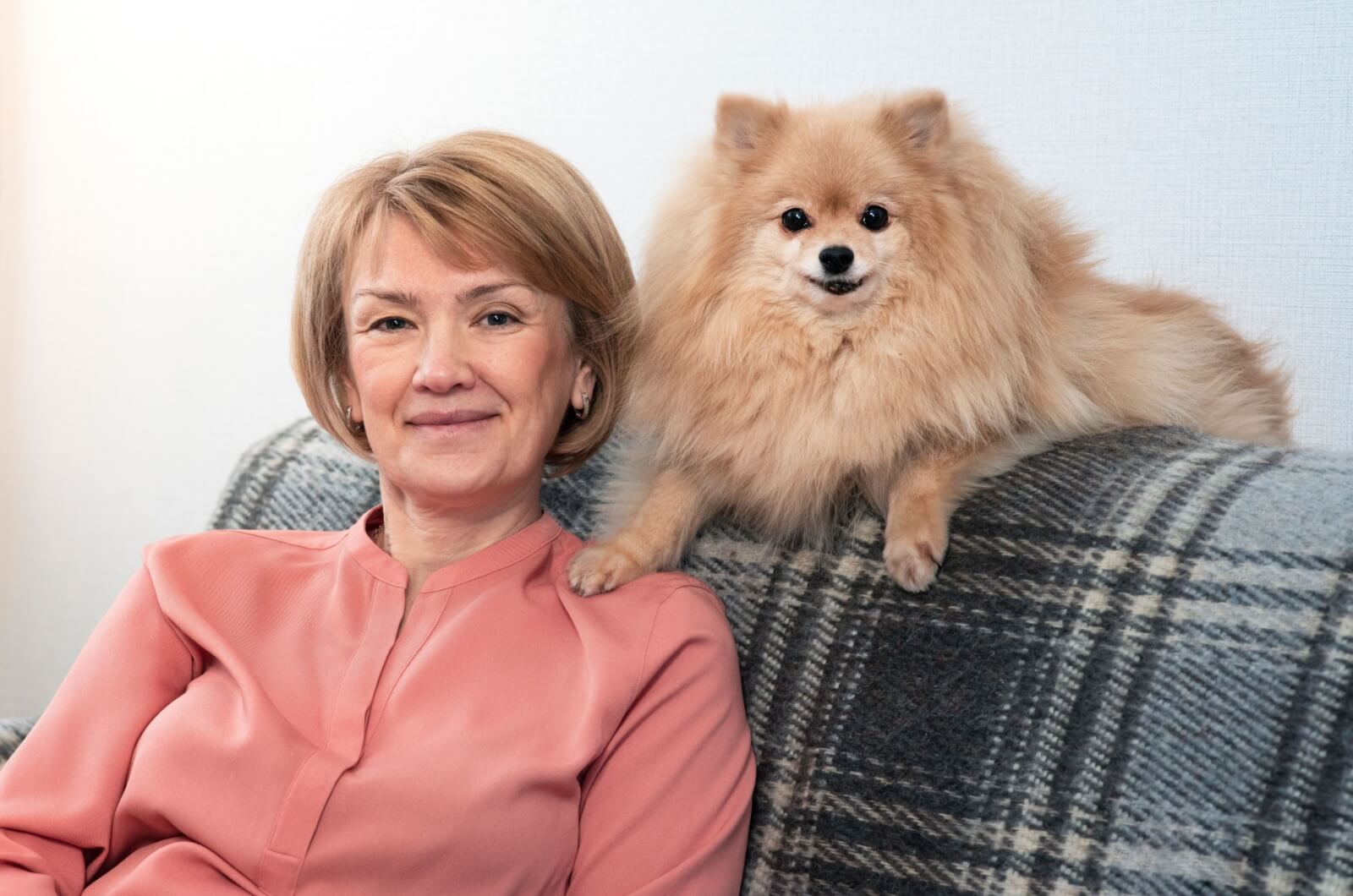 A senior woman sitting on a couch with a Pomeranian dog sitting on the back of the couch