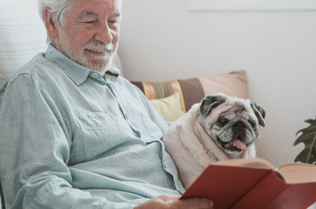 An older man sitting on a couch holding a book with a pug next to him