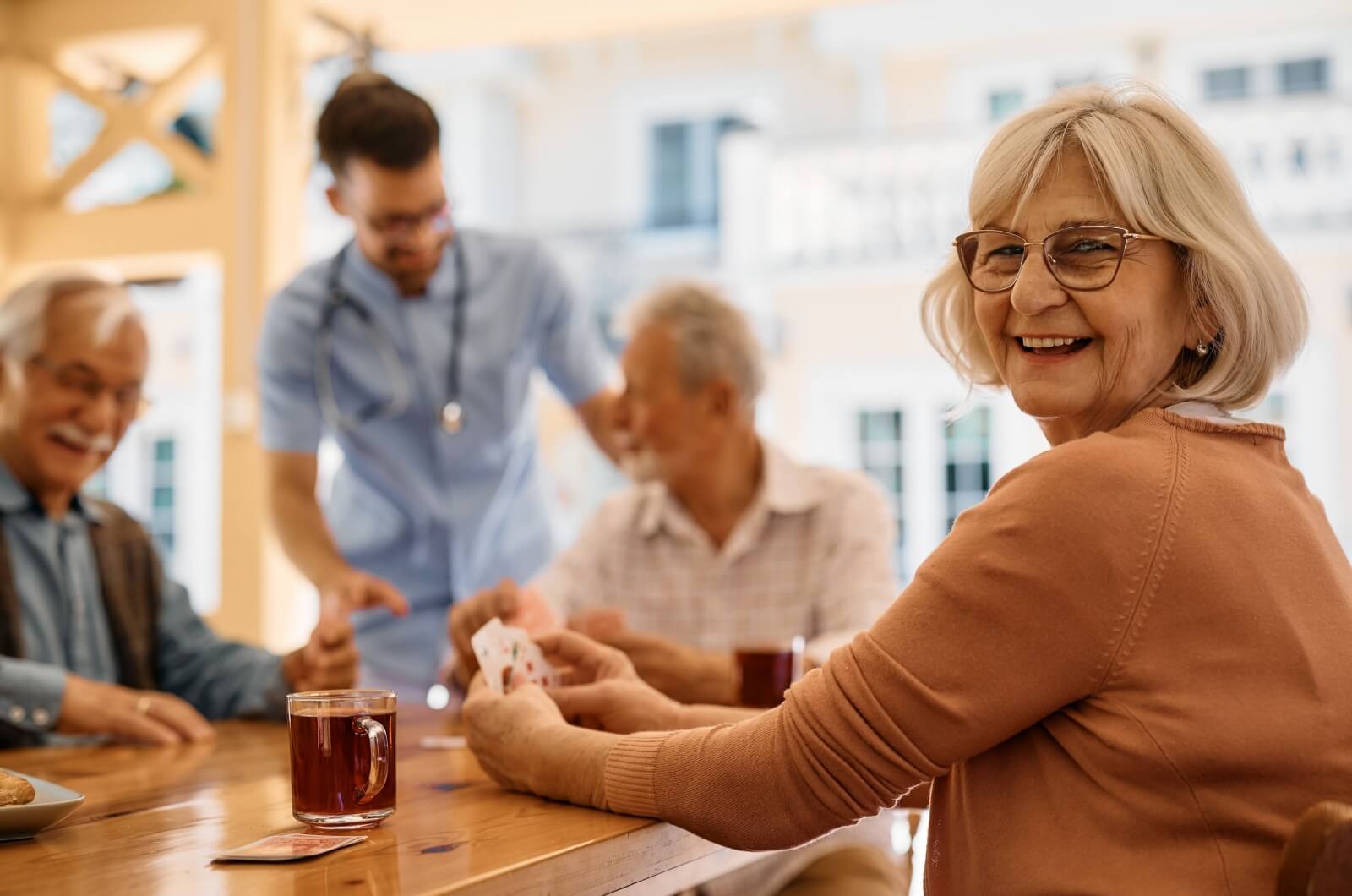 A group of smiling seniors in an assisted living community playing cards together while a doctor checks in on them.