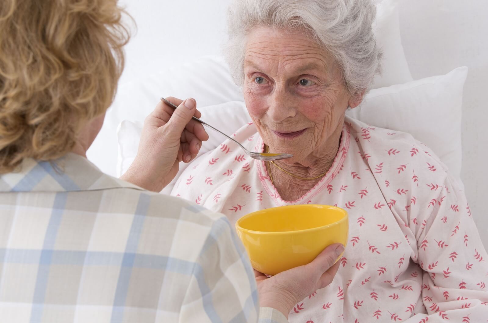 A woman holds a spoon and a bowl of soup, attentively feeding an elderly lady who is experiencing a decrease in appetite.