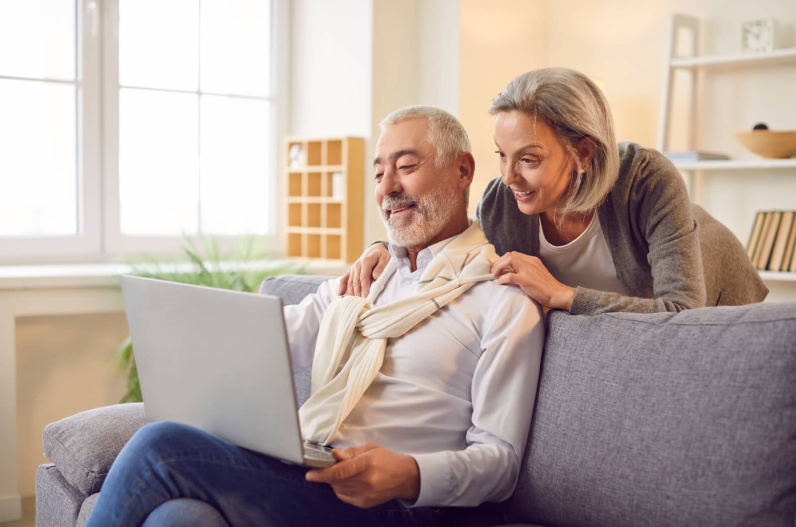 An older man sitting on a couch with his spouse leaning over his shoulder looking at assisted living forms on a laptop.