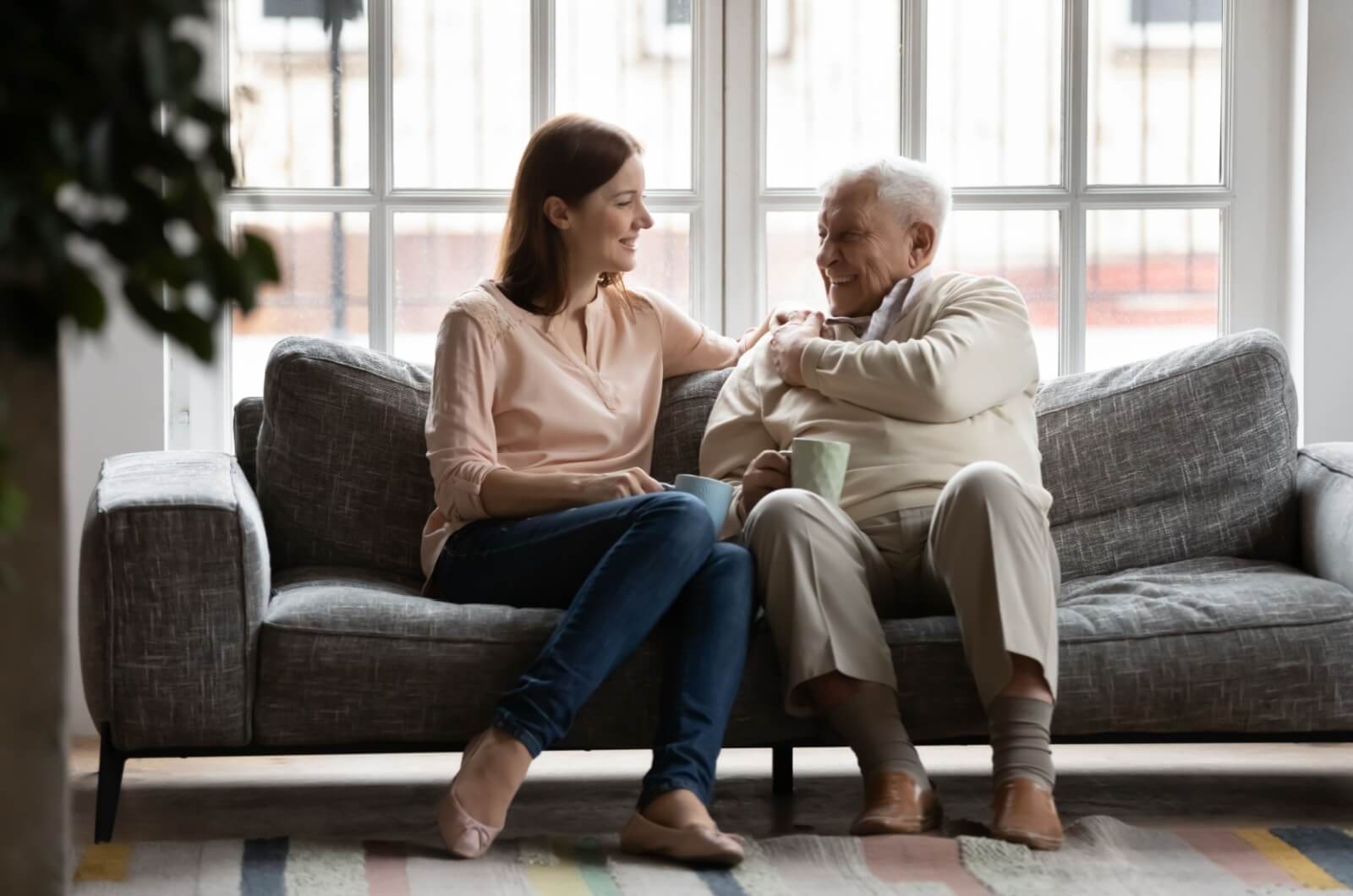 An adult child sitting on the couch with their parent and smiling while discussing memory care.