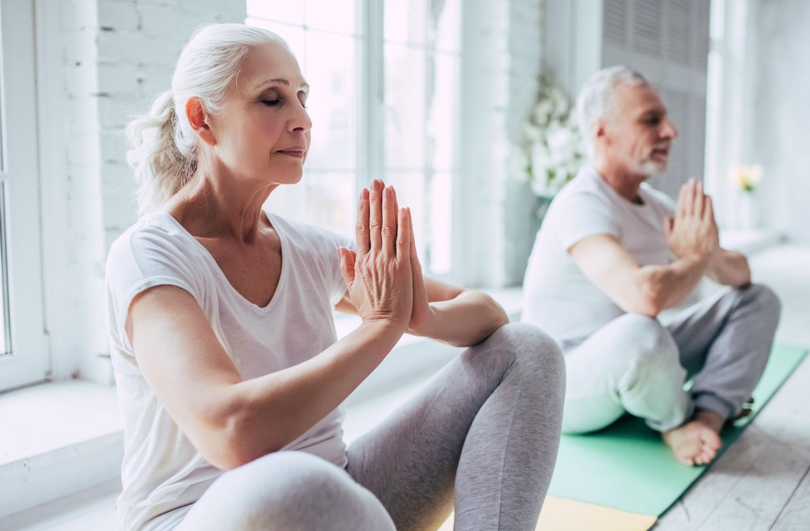 An older couple sitting crossed legged on yoga mats with their eyes closed and hands folded.