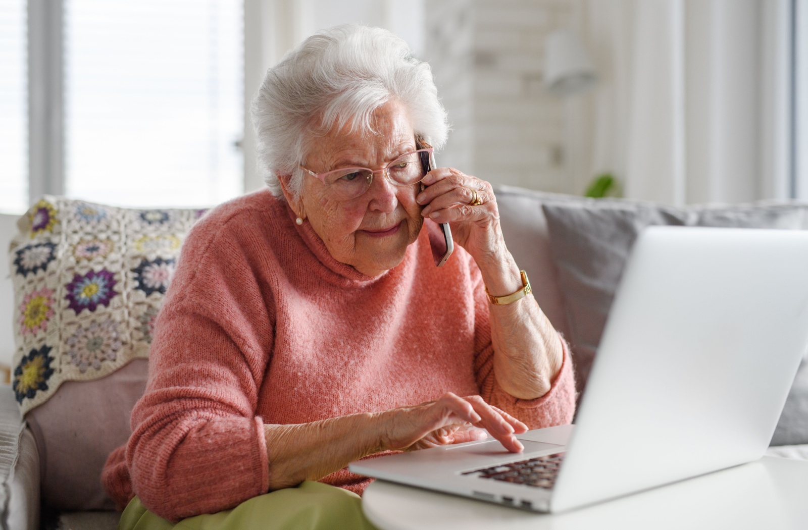 An older adult holding a phone in one hand while browsing on a laptop with the other hand.