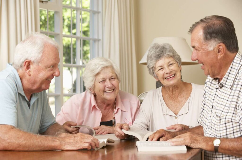 4 older adults around a kitchen table smiling with open books during their book club meeting.
