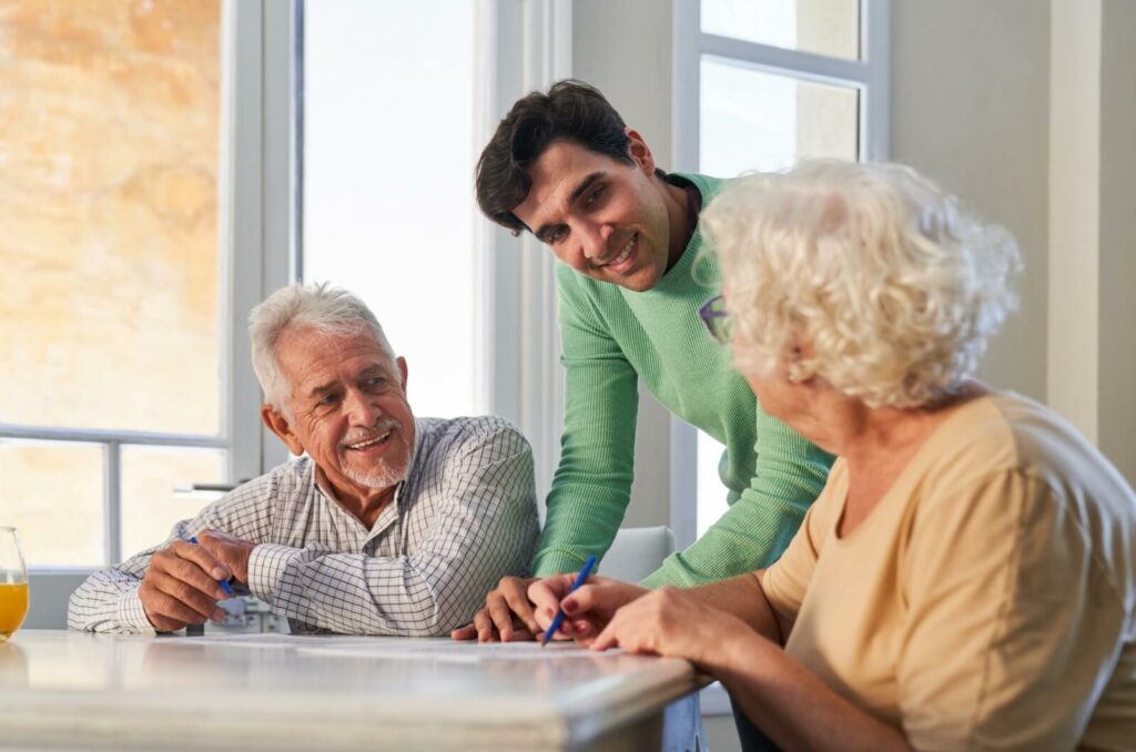 A caregiver smiling as they help 2 memory care residents sitting at a table work on a memory care activity.